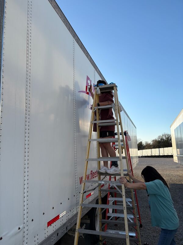 Two-person crew with a tall ladder lettering the side of a white semi trailer